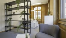 Office with wooden paneled walls, white ceilings, open layout, floor-to-ceiling windows, shelves, black chair, white desk, potted plants