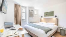 Spacious bedroom with large windows, beige curtains, white walls, hardwood flooring, and modern furniture including a bed, desk, and shelving unit. The room also features artwork on the walls and a laptop on the desk, suggesting it may be used as a worksp