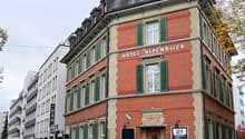 A multi-story hotel with red brick walls and green shutters, named Hotel Alpenblick, located on a street with parked cars and trees, next to another white building with many windows.