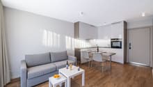 Open space living room with kitchen, dining area, and seating area with a gray couch, white tables, wooden floor, white kitchen cabinets, and stainless steel countertops.