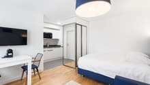 The image shows a modern and minimalist studio apartment. The room features a white and blue color scheme, with a large bed, a desk with a chair, and a kitchenette area with a microwave and other appliances. The room has hardwood floors and a large window