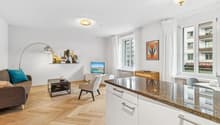 open living space with kitchen, large couch with cushions, armchair, coffee table, tv stand with flat screen tv, floor lamp, wooden parquet flooring, kitchen island with granite countertop, dining area with dining table and chairs, picture frames on the w