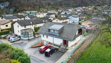 Aerial view of a small town or village nestled in a mountainous, forested landscape. The image shows a cluster of residential buildings with tiled roofs, some with balconies or terraces. There are several parked cars visible in the foreground, indicating