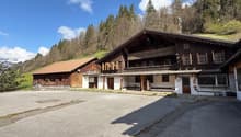 A large wooden chalet-style building with a parking area in front, surrounded by a forested mountain landscape.