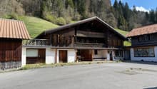 A traditional wooden chalet-style building with a sloped roof, surrounded by a grassy area and trees. The building has a covered porch and several garage doors or parking spaces on the ground floor.