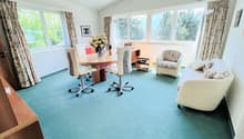 Spacious home office with large windows, wooden desk, office chairs, floral armchair, and a sofa. The room has a blue carpeted floor and appears to have access to a balcony or terrace.