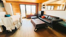 Spacious living room with wooden walls and ceiling, large windows, leather sofa, coffee table, and a balcony or terrace visible.