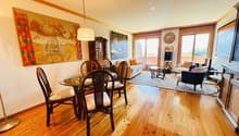 Wooden flooring, round dining table with chairs, hanging lamp, paintings on the wall, glass sliding doors, couch with pillows, side table with lamp, coffee table, rug on the floor, cabinet