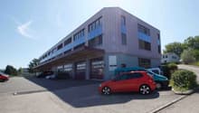 A multi-story commercial building with a modern, angular design featuring large windows. The building has a parking lot in front with several cars parked. The surroundings include some trees and greenery.