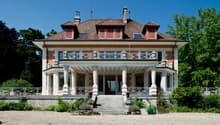 Raised ground floor, tiled roof, brown brick, white exterior, large balcony with railings, staircases, stone porch, windows, plants