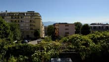 Outdoor view of a row of multi-story buildings with mountains in the distance, a street, and a car parked in the front. Some plants are growing over a small fence.
