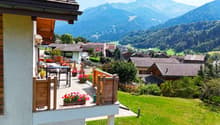 Apartment with balcony, mountain view, wooden railing, outdoor seating with table and chairs, flower pots, green lawn, several buildings and a mountain range in the background.