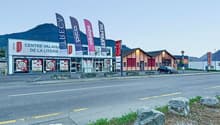 A commercial building with a car parked outside, flags on poles, large windows, red doors, mountains in the background.