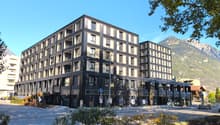Modern 6-story building, black exterior with white accents, several balconies visible, storefronts on the ground floor, surrounded by trees and shrubs