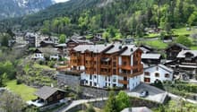 The image shows a picturesque alpine village nestled in a lush, mountainous landscape. The buildings are primarily made of wood and stone, with traditional alpine architecture featuring sloped roofs and balconies. The village is surrounded by dense forest