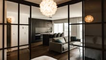 modern living room with glass walls, wooden floor, gray couch, coffee table, kitchen with island, dining table, balcony