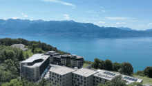 Modern building complex with multiple buildings, solar panels, surrounded by trees, overlooking a lake and mountains
