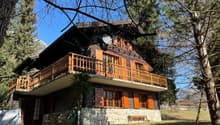 Two-story wooden chalet-style house with a balcony, surrounded by trees and a grassy area
