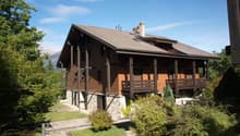 A two-story chalet with a brown wooden exterior, gray roof, wooden front porch, multiple windows, surrounded by a grassy area with various plants and trees.