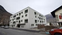 Multi-story apartment building with gray exterior, balconies, and windows. The building is situated in a mountainous area with other buildings and vehicles visible in the surroundings.