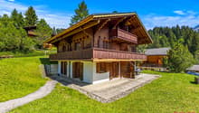 2-story wooden chalet-style house with a balcony, surrounded by a grassy lawn and pine trees in a mountainous, forested area