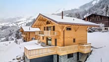 2-story wooden chalet-style house with a stone base, large balconies, and a garage. The house is surrounded by snow-covered mountains and trees.