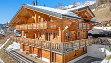 Two-story wooden chalet with a sloped roof, covered balconies, and a snowy mountain backdrop