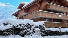 A multi-story wooden chalet-style building with snow-covered roofs and balconies, surrounded by snow-covered trees and a snowy landscape.