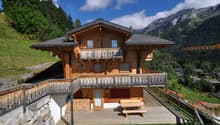 2-story wooden chalet-style house with a balcony, wooden deck, and parking space. The house is surrounded by lush green trees and mountains in the background.