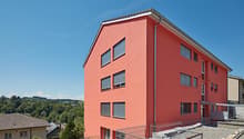 Modern apartment building, red exterior, multiple windows, metal railings, gravel pathway, nearby car