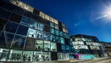 A modern, multi-story office building with a glass facade and visible parking spaces in front. The building has a sleek, contemporary design with large windows allowing natural light to enter the interior spaces.