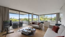 large sliding glass doors leading to a balcony, hardwood floors, large windows with scenic view, modern grey sofa, coffee table, flatscreen TV on stand, minimalist decor