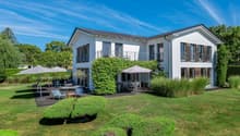Two-story white house with balconies, surrounded by a well-maintained garden with trees and shrubs. There is a patio area with outdoor furniture in the backyard. The house has a tiled roof and large windows.