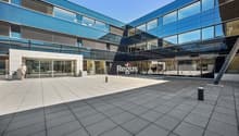 Modern glass and steel commercial building with the Regus logo prominently displayed. The building has a large open plaza area with paved flooring in front of the main entrance.