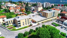 Multi-story building with a flat roof, situated near a road, surrounded by greenery and buildings
