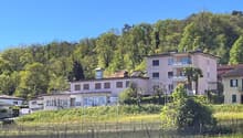 A multi-story residential building with balconies, surrounded by trees and greenery in a scenic, hilly landscape.