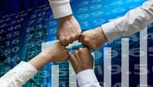 The image shows the hands of several people coming together in a fist bump, symbolizing teamwork and collaboration. The background features a cityscape with skyscrapers and financial data, suggesting a business or corporate setting.