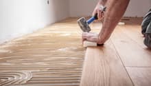 Hardwood floor being installed on concrete base, person using hammer, wall with white paint, grooves for installation