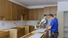 Unfinished kitchen, wooden cabinets, countertop, electrical outlet on wall, sawing machine, open door, man using mask, hammer on belt
