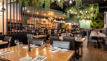 Upscale restaurant with a cozy and modern interior design. The space features wooden tables, leather chairs, and a bar area with a large selection of wines and liquors. The ceiling is adorned with hanging greenery and string lights, creating a warm and in