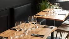Wooden dining table set with wine glasses, cutlery, and a plant decoration. Dark walls and furniture create a cozy atmosphere. The table is set for a dining experience, with the visible features including a dishwasher, suggesting this is a restaurant sett