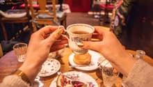 The image shows a person's hands holding a floral-patterned ceramic mug filled with a hot beverage, likely tea or coffee, on a table with other dishes and plates. The background suggests a cozy cafe or restaurant setting with wooden furniture and decor.