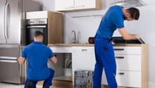 modern kitchen with stainless steel appliances, white cabinets, tiled flooring, two men in blue uniforms, toolbox and tools on the floor