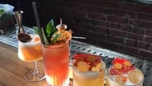The image shows a bar setting with various cocktail glasses and bottles on display. There are three cocktail drinks on the wooden bar top, each garnished with different fruits and herbs.