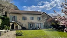 Two-story stone house with green shutters, surrounded by a well-maintained garden with a cobblestone driveway, flowering trees, and outdoor seating area
