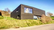 Modern house, black exterior, large windows, elevated position, surrounded by greenery