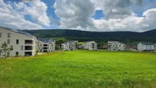 A modern residential complex with multiple apartment buildings surrounded by a large grassy field and hills in the background. The buildings have balconies and are painted in light colors.