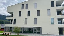 Multi-story apartment building with a modern, minimalist design. The building has large windows, balconies, and a paved outdoor area with a picnic table. The exterior is painted in a light, neutral color with black window frames and railings.