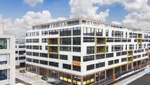 A modern, multi-story commercial building with a large glass facade and yellow accents. The building appears to have a parking area in front of it.