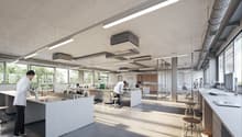 The image shows a modern, open-plan office space with a kitchen area. The space features large windows, allowing natural light to flood in. There are various workstations with desks and chairs, as well as a kitchen counter with what appears to be a dishwa